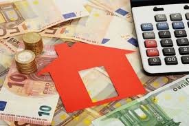 Dinero en efectivo, monedas y una calculadora junto a una figura roja de casa, simbolizando la financiación inmobiliaria o inversiones en bienes raíces.