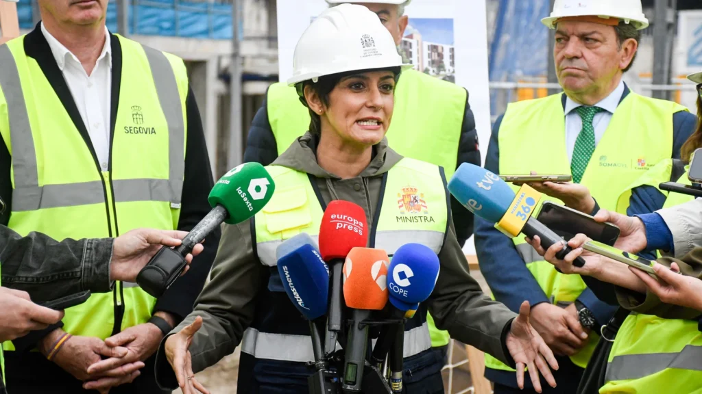 Ministra hablando ante la prensa en una obra de construcción, rodeada de periodistas con micrófonos y personal con chaleco de seguridad.