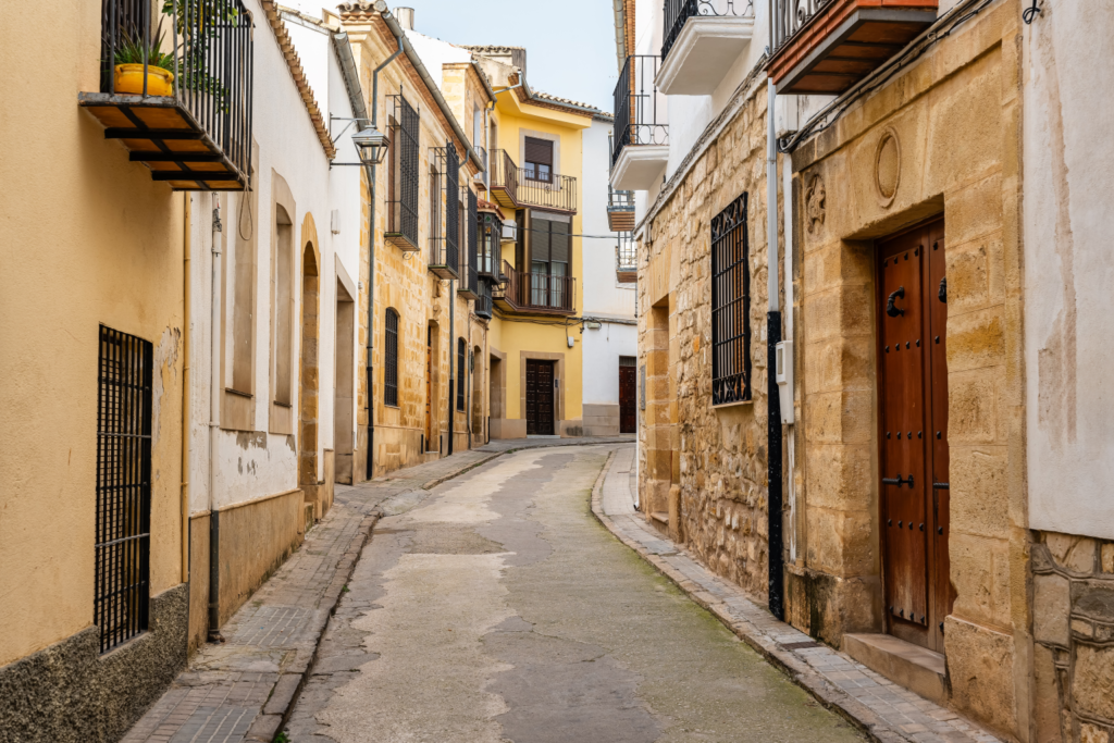 Teresa Gestora Alquiler Vacacional. Calle empedrada de pueblo en España con edificios antiguos de piedra y balcones. Cobblestone village street in Spain with old stone buildings and balconies.
